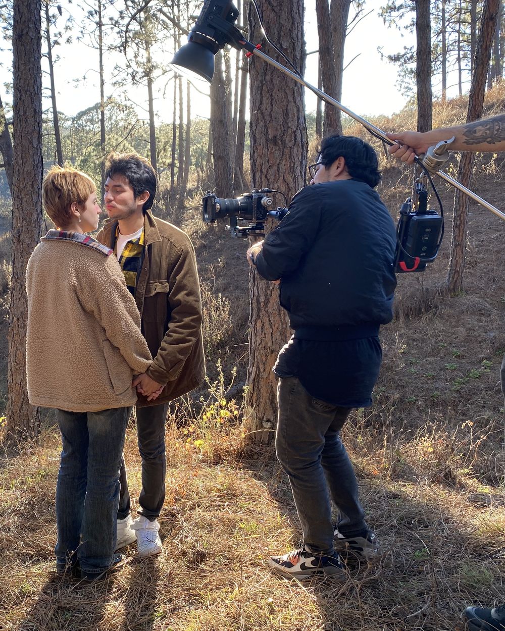 Equipo de rodaje filmando a una pareja en un bosque de pinos, con cámara al hombro y una luz en pértiga